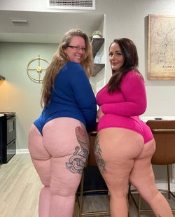 Two curvy women in form-fitting bodysuits pose back-to-back in a kitchen, showcasing tattoos on their thighs and