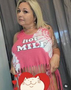 Blonde woman in a pink tie-dye top with 'hot MILF' text, holding the hem to reveal midriff, standing against a gray