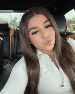 Brunette woman with long hair in a white zip-up top, making a playful face while sitting in a car with black and tan