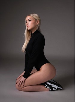 Blonde woman in black long-sleeve bodysuit kneeling on gray background, wearing black and white sneakers.