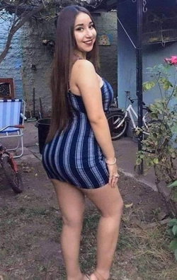 A woman with long dark hair in a blue and white striped dress poses outdoors in a garden setting, smiling at the camera.