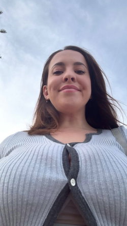 A woman with long brown hair smiles softly in a low-angle selfie, wearing a gray ribbed cardigan outdoors under a