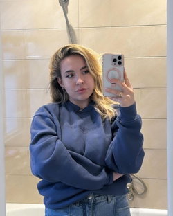 Blonde woman in a blue sweatshirt and jeans takes a mirror selfie in a tiled bathroom, arms crossed.