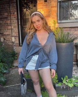 A blonde woman in a sheer blue shirt and white shorts poses outdoors near a brick building with plants.