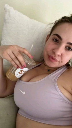 A woman with dark hair lies on a pillow, wearing a lavender sports bra and holding a drink, looking at the camera.