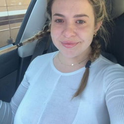 A young woman with braided hair smiles in a car wearing a light blue top.