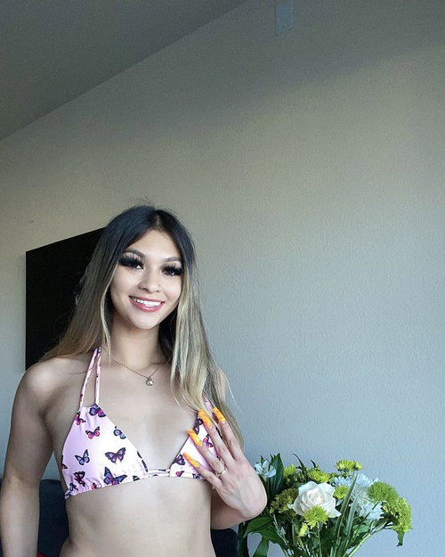 A smiling woman with long ombre hair wears a butterfly-print bikini top, standing indoors near a bouquet of flowers.