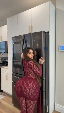 Curvy brunette in sheer red lace bodysuit leans against fridge in modern kitchen.