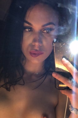 Brunette woman with dark hair and makeup takes a mirror selfie, bare-chested, holding phone with visible flash.
