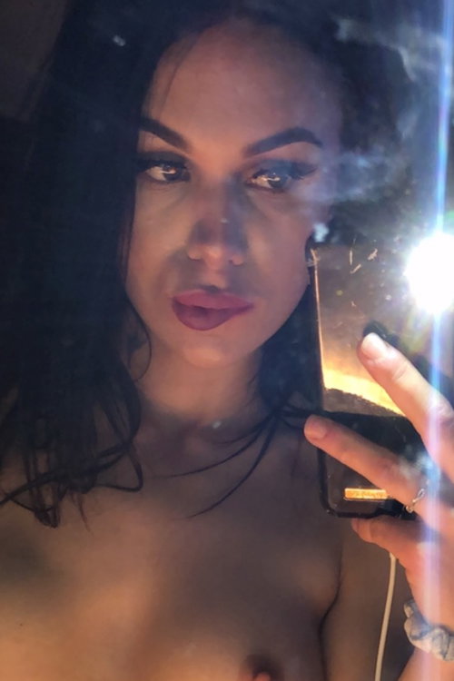 Brunette woman with dark hair and makeup takes a mirror selfie, bare-chested, holding phone with visible flash.