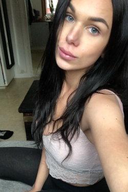 A woman with long dark hair and blue eyes wears a light pink lace crop top and black pants, sitting indoors in a casual