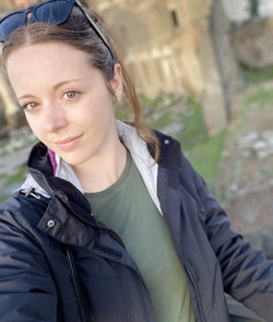 A young woman with brown hair and green eyes wears a dark jacket over a green tee, smiling softly in an outdoor rocky