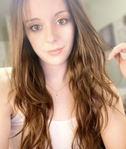 A woman with long wavy brown hair and a white tank top smiles softly at the camera in a softly lit indoor setting.