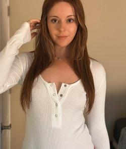 Redheaded woman in a white ribbed long-sleeve top with buttons, posing indoors with hand in hair.