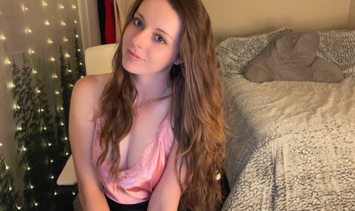 Curvy brunette in pink lace camisole and black shorts smiles on bed with fairy lights.