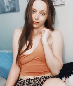 Brunette woman with long hair in a brown crop top and leopard-print skirt, sitting on a couch indoors.