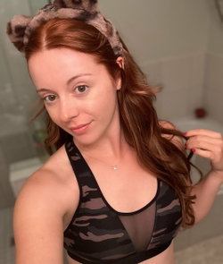 Woman with long brown hair in a messy bun, wearing a black sports bra, taking a selfie in a bathroom.