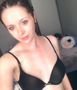 A woman with brunette hair in a black bra poses in a bedroom.