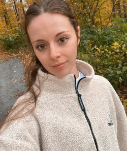 A woman with brown hair in a ponytail wears a cream fleece jacket outdoors in a wooded area with autumn foliage.