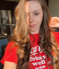 Woman with long wavy brown hair wearing a red graphic t-shirt, taking a selfie in a kitchen setting.