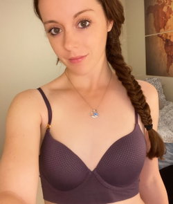 Woman with long brown braided hair wearing a purple textured bra, taking a selfie indoors with soft lighting.
