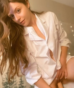 A woman with long brown hair wears an unbuttoned white satin shirt, sitting indoors with soft lighting and string