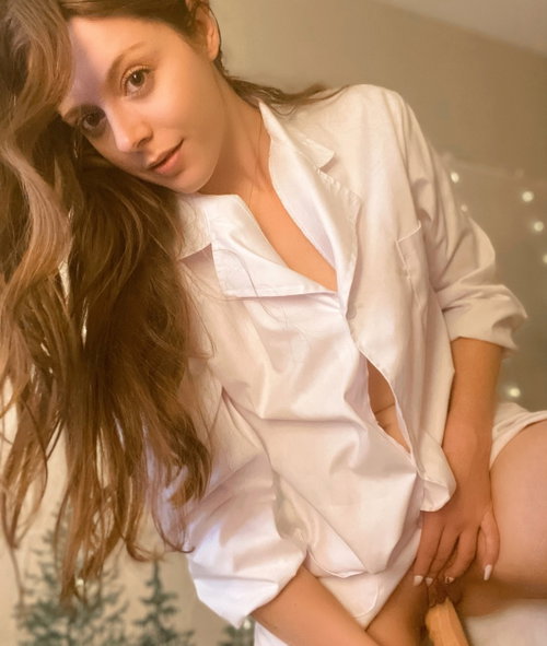 A woman with long brown hair wears an unbuttoned white satin shirt, sitting indoors with soft lighting and string