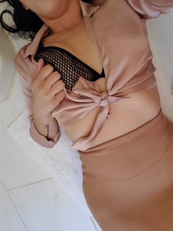 Woman with dark hair wearing a sheer pink top over a black mesh bra, posing on a white rug with a tattoo visible on her