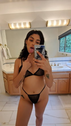 Brunette woman in black mesh lingerie takes a mirror selfie in a bathroom, sticking out her tongue and making a hand