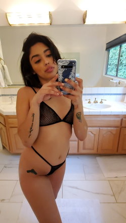 Brunette woman in black mesh lingerie takes mirror selfie in bathroom, showing tattoos on arms and hip.