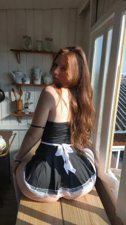 A woman with long hair in a black and white maid outfit sits on a windowsill in a kitchen.