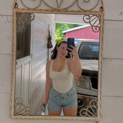 A curvy woman with dark hair takes a mirror selfie in a white tank top and denim shorts, holding a water bottle