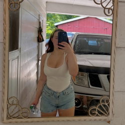 Curvy woman in white tank top and denim shorts takes mirror selfie outdoors, holding water bottle, with white truck and