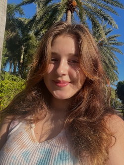 A woman with long brown hair smiles softly in a sunny outdoor setting with palm trees behind her, wearing a