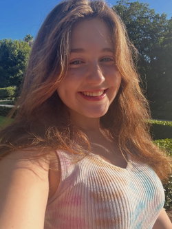 A smiling woman with long brown hair wearing a pastel ribbed tank top, outdoors in a sunny park setting.