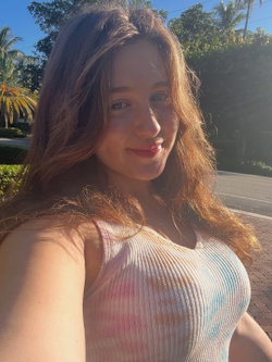 A woman with long brown hair smiles in a colorful ribbed tank top, outdoors on a sunny day with palm trees in the
