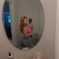 A woman with red hair takes a mirror selfie in a pink bikini top and denim shorts in a bathroom.