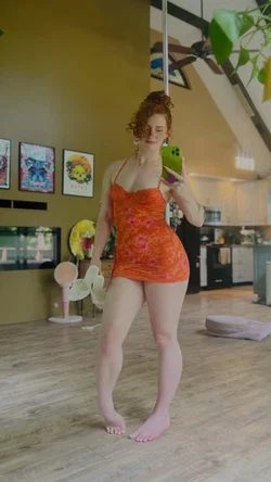 Redhead woman in orange dress taking a selfie in a bright kitchen.