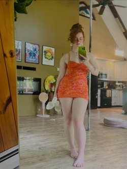 Redhead woman in a coral dress taking a selfie in a well-lit room with wooden floors and a kitchen in the background.