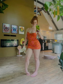 Redhead woman in orange dress taking a selfie in a bright kitchen.