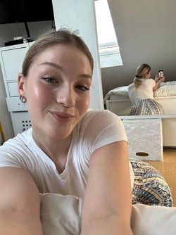A young woman in a white t-shirt takes a selfie in a bedroom.