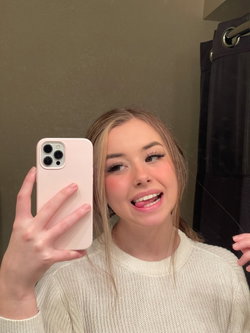 Blonde woman in a cream knit sweater takes a mirror selfie, smiling playfully with makeup and styled hair in a bathroom
