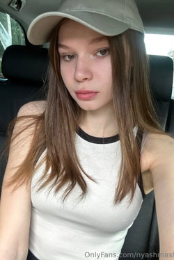 Woman with long brown hair wearing a white tank top and gray cap, sitting in a car, looking at the camera.