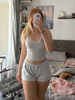 Blonde woman in gray crop top and shorts takes mirror selfie in bedroom with bed and desk visible.