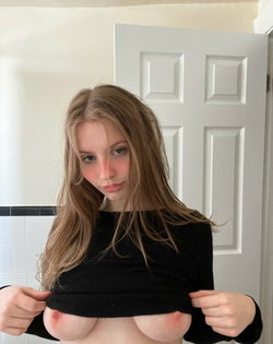 A woman with long light brown hair lifts her black top to expose her bare breasts, standing in front of a white door
