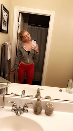 Blonde woman in a gray cropped hoodie and red leggings takes a mirror selfie in a bathroom, sticking her tongue out.