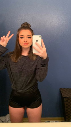 A young woman with brown hair in a bun takes a mirror selfie, wearing a dark long-sleeve top and black shorts, making a