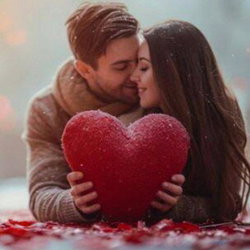 A couple embraces closely in the snow, holding a large red heart, surrounded by scattered rose petals.