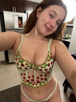 A woman with brown hair in a strawberry-patterned lingerie set poses for a selfie in a kitchen.