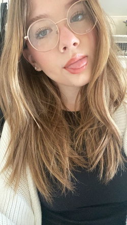 A young woman with long blonde hair and round glasses sticks her tongue out playfully, wearing a black top under a